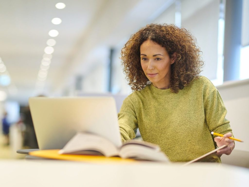 A woman in a green sweater looks at her laptop while sitting in the library.